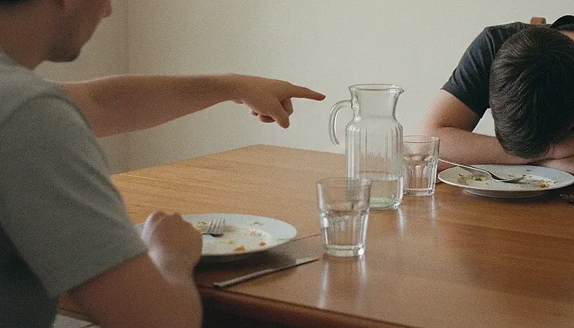 A hand extended across a dining table with the index finger pointing, the speaker cut off above the shoulders by the top edge of the frame, while the other person rests their head on their folded arms beside an unfinished plate.